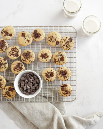 Low FODMAP chocolate chip pancake mini muffins on a cooling rack with a small cup of almond milk and a bowl of chocolate chips.
