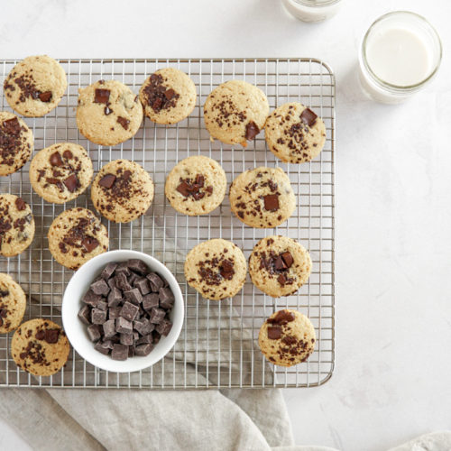 Low FODMAP chocolate chip pancake mini muffins on a cooling rack with a small cup of almond milk and a bowl of chocolate chips.