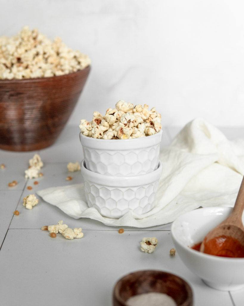 A small bowl of Low FODMAP Buffalo Ranch Popcorn, with a large bowl in the back and a small side of hot sauce and fine sea salt for decoration.