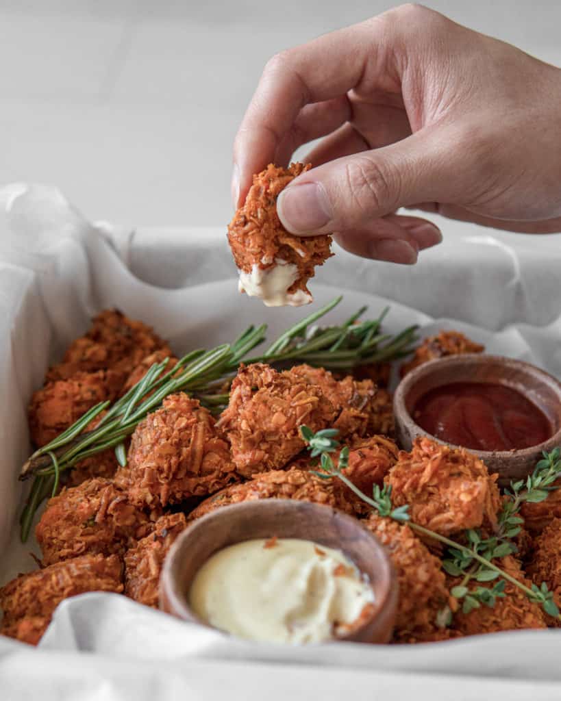 Crunchy Low FODMAP sweet potato tots piled in a tray with two sauces bowls of Lemon Aioli and Ketchup. Rosemary and Thyme are used as decorative garnish. A hand is grabbing one tot that has been dunked in the Lemon Aioli.