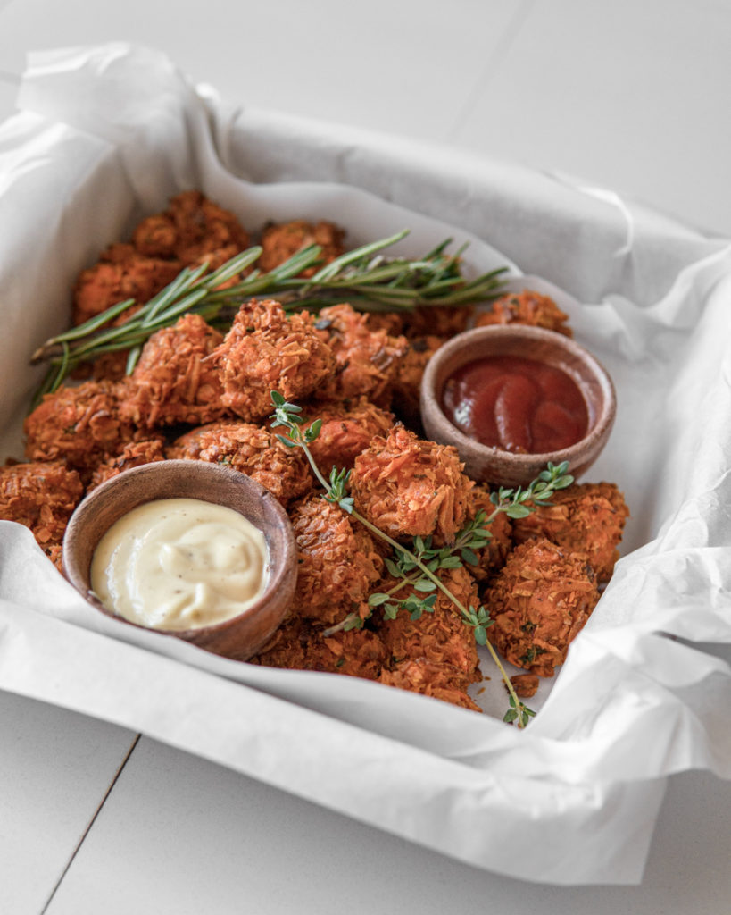 Crunchy Low FODMAP sweet potato tots piled in a tray with two sauces bowls of Lemon Aioli and Ketchup. Rosemary and Thyme are used as decorative garnish.